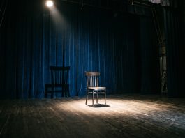 A single spotlight on an empty chair on a dark stage, suggesting a sci-fi horror theme.