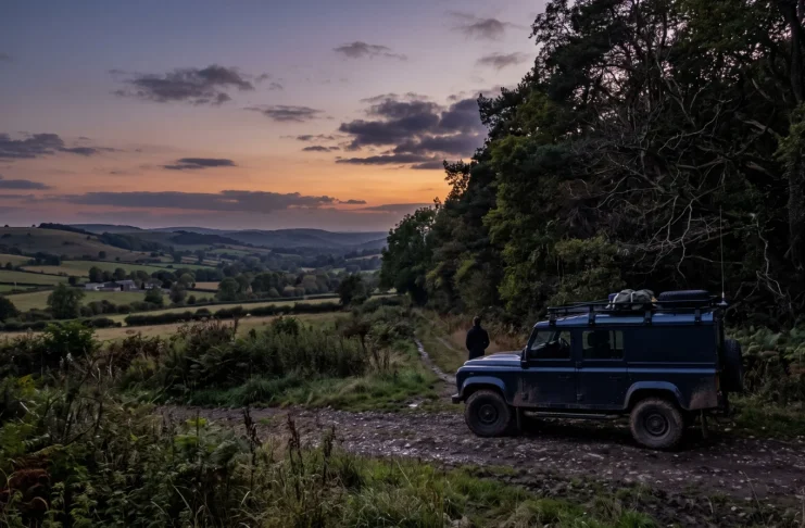 A car parked near a dark forest edge in the British countryside.